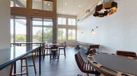 This contemporary amenity lounge is designed to impress, combining stylish industrial elements with warm, inviting finishes. The space is defined by its dramatically soaring double-height ceilings, exposed ductwork, and expansive floor-to-ceiling windows and glass doors that flood the interior with natural light and frame lush tree canopy views beyond an adjacent outdoor balcony. Dark slate-toned tile flooring runs throughout the main gathering area, complementing the neutral warm-beige walls and modern wall sconces.

The main lounge area is furnished with a mix of high-top bistro tables with bar stools, a long glass-top counter with seating, and a dedicated game zone featuring a full-size shuffleboard table. A round poker table surrounded by leather swivel chairs anchors a separate game room nook, accented by a bold geometric pendant light and a wall-mounted flat-screen TV.

A well-appointed wet bar area adds functional elegance, showcasing deep navy-blue cabinetry, a white quartz countertop, a sink, under-counter refrigeration units, and richly textured dark subway tile extending floor to ceiling. A framed Scrabble board adds a playful decorative touch. Together, these zones create a versatile, entertainment-focused space suited for casual gatherings and organized social events alike.