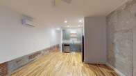 Located at 1200 S 2nd Street in Wilmington, North Carolina, this two-floor space offers a well-considered layout with a blend of modern updates and character-rich details. The warm, light-toned hardwood floors run throughout, lending a welcoming continuity across both levels and complementing the bright, neutral walls.

The kitchen area is thoughtfully designed with sage-toned cabinetry, open shelving, stainless steel appliances including a dishwasher and refrigerator, and a functional sink — all set within a compact yet efficient footprint. Recessed lighting illuminates the space cleanly, while a wall-mounted mini-split unit provides climate control. The open-plan lower level connects the kitchen to a generous living area, creating a spacious and versatile environment suitable for a variety of uses.

The dollhouse view reveals the structural character of the upper floor, where exposed wood framing and raw architectural elements give the space a distinct, industrial edge. Natural light filters in through windows on the upper level, enhancing the airy feel of the interior. The two-floor configuration provides clear separation between areas, offering functional flexibility throughout. Overall, the space balances contemporary finishes with raw, authentic architectural details in a central Wilmington location.