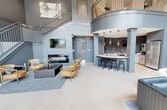 This 1,060-square-foot amenity lounge is a single-floor space designed with a contemporary aesthetic and striking architectural details. The centerpiece of the room is its dramatic double-height ceiling, which floods the interior with natural light through expansive floor-to-ceiling windows that offer views of an outdoor pool courtyard. The light-toned large-format tile flooring anchors the open layout, while the walls are finished in a sophisticated slate gray palette throughout.

The space is organized into several distinct functional zones. A primary lounge area features a curated mix of tan leather accent chairs, a modern sofa, and a low-profile coffee table arranged on a soft blue-gray area rug, complemented by a wall-mounted television and built-in cabinetry. A secondary seating arrangement with rounded, contemporary chairs provides additional gathering space near the windows. A dining area with a solid wood table and chairs sits adjacent to a full kitchenette bar, complete with a dark countertop island, bar stools, and pendant lighting.

A staircase with steel railings leads to a curved mezzanine balcony overhead, adding visual depth and architectural interest. Lush indoor plants are positioned throughout the space, enhancing the welcoming ambiance. Recessed and pendant lighting complete the polished, upscale atmosphere of this well-appointed communal space.