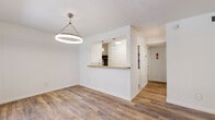 This well-appointed single-floor unit offers three bedrooms and one bathroom within a thoughtfully organized layout. Wood-look plank flooring runs throughout the entire space, providing a warm and cohesive aesthetic from room to room.

The combined living and dining area serves as the heart of the home, featuring a striking circular pendant light fixture and a large window that invites natural light and offers a view to the exterior. The open-concept kitchen connects to the living area via a pass-through counter with a warm wood-toned surface, complemented by decorative pendant lights above and a subway tile backsplash. A built-in shelving nook adds a functional accent to the kitchen area.

The primary bedroom is a comfortable, well-proportioned room equipped with a ceiling fan and light fixture, and benefits from its own dedicated closet space. The additional two bedrooms are clean and bright, each with wood-look flooring. One bedroom notably includes two separate closets, providing generous storage capacity.

The bathroom is finished with classic white hexagonal mosaic floor tile, a full tub and shower combination, and a window that brings in natural ventilation and light. The overall palette throughout the unit is clean and neutral, emphasizing white walls and modern brushed nickel hardware, creating a fresh and move-in-ready environment.