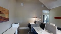 This single-floor workspace offers a functional and modern environment suited for professional or collaborative use. The room's most distinctive architectural feature is its vaulted ceiling, which rises dramatically along a sloped roofline, adding a sense of openness and volume that extends well beyond what the floor area alone might suggest.

The workspace is equipped with multiple computer workstations arranged along clean-lined white desks, each fitted with acoustic privacy panels in neutral grey tones to support focused individual work. Ergonomic task chairs in dark upholstery complement the contemporary aesthetic. A warm-toned desk lamp provides supplementary ambient lighting, contributing to a calm and productive atmosphere.

Built-in in-wall speakers are mounted at upper wall positions, suggesting integrated audio capabilities throughout the space. A security camera is also visible near the ceiling junction, indicating the space is equipped for monitoring. Neutral taupe-toned walls and dark flooring create a cohesive, understated palette throughout.

A glimpse through an adjacent opening reveals a connected area with glazed partitioning and cabinetry, suggesting additional functional zones nearby. A printer station is positioned conveniently along one wall, rounding out the practical, well-considered layout of this workspace.