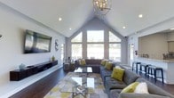 This single-story space makes a striking impression with its dramatic vaulted cathedral ceiling, which rises to a sharp peak and creates a sense of generous vertical space throughout the main living area. The architectural design is anchored by a large, symmetrical window arrangement at the far wall, following the roofline's triangular profile and flooding the interior with natural light while offering open views of the surrounding landscape.

The living area is generously proportioned, accommodating a large L-shaped sectional sofa in charcoal gray, accented with mustard yellow and white throw pillows that add a contemporary pop of color. A geometric-patterned area rug in gray and yellow tones defines the seating zone, complemented by glass-top coffee tables with metallic bases. A wall-mounted flat-screen television is positioned opposite the seating arrangement, with a sleek media console below.

Overhead, a modern gold-toned geometric chandelier serves as a focal point, adding warmth to the otherwise cool gray palette. Recessed lighting supplements the natural light. Dark hardwood flooring runs throughout, providing rich contrast to the light gray walls and white ceiling, resulting in an inviting, well-balanced ambiance.