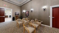Located at 3980 S Jackson Dr in Independence, MO, this expansive single-floor facility offers a generously sized, thoughtfully designed interior with a welcoming and upscale ambiance. The main gathering area is an impressive, open space anchored by soaring ceilings, skylights that flood the room with natural light, and a grand chandelier. Wood flooring, ornate area rugs, and classic furnishings — including a grand piano and organ — lend a refined, elegant character to the space. Wide, well-lit hallways connect the various areas, featuring patterned carpet, decorative artwork, wall sconces, and a wood-trimmed elevator, all contributing to a warm and navigable layout. The game room is outfitted with seating arranged around a projection screen, wall-mounted sconces, and artwork, creating a comfortable space for entertainment and gatherings. A dedicated activity or classroom area provides tables and chairs with ample lighting, suitable for programming and group activities. A cozy nook with rich terracotta-toned walls, a decorative column, a wooden desk with computer access, and an armchair serves as a quiet retreat or workstation. Throughout the facility, consistent use of warm tones, tasteful décor, and quality finishes creates a cohesive, inviting environment.