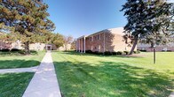 This single-floor community space is housed within a classic two-story brick building surrounded by expansive, manicured lawns, towering mature trees, and neatly trimmed shrubbery. A clean concrete walkway leads visitors to the entrance, creating an inviting first impression under open blue skies.

Inside, the space is characterized by white shiplap-style wall paneling that lends a clean, cottage-inspired aesthetic throughout. The main gathering area is anchored by a striking floor-to-ceiling stone fireplace, a true standout feature that adds rustic warmth and visual character to the room. Exposed ceiling beams and a ceiling fan overhead contribute to the relaxed, welcoming ambiance.

The lounge area offers generous seating with plush sofas and armchairs arranged around a central coffee table, ideal for comfortable conversations or casual meetings. A dedicated workspace with a desk and office chairs is situated nearby, seamlessly integrating a functional leasing or administrative area within the open-concept layout.

Adjacent to the main lounge, a dining area features a glass-top round table with wood chairs and direct access to the outdoors through a sliding glass door, allowing natural light to fill the space. Decorative shelving with curated accents adds a personal, homey touch. Patterned carpeting, arched window treatments, and tasteful artwork complete the warm, community-centered atmosphere of this well-appointed space.
