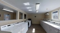 This single-floor laundry facility is designed with practicality and efficiency in mind. The room features a generous layout with multiple top-load washing machines lining one wall and a row of dryers positioned along the opposite side, providing ample capacity for shared use. The warm beige walls are complemented by a neutral speckled floor, lending the space a clean and utilitarian aesthetic. Bright fluorescent lighting fixtures mounted on the white ceiling ensure the room is well-lit throughout. A row of mirrors is mounted along one wall, adding a sense of openness and depth to the space. Crown molding provides a subtle architectural detail that ties the room together. Ventilation is addressed with ceiling vents, maintaining airflow and comfort during use. The space also includes what appears to be a folding or utility counter area, offering a convenient surface for sorting and organizing laundry. Overall, the facility is thoughtfully arranged to maximize usability while maintaining a tidy and welcoming environment suited to its functional purpose.