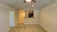 Located in Richardson, Texas, this single-floor residential space offers a thoughtfully arranged open-concept layout designed for comfortable everyday living. The living area is finished with wall-to-wall carpeting that adds warmth underfoot, and is equipped with a ceiling fan fitted with an integrated light fixture to ensure year-round comfort and ambient lighting. Recessed lighting is incorporated throughout, providing consistent, even illumination across the space. A defining architectural feature is the raised pass-through peninsula that connects the living area to the kitchen, creating a natural division between spaces while maintaining an open and airy feel. The kitchen is outfitted with dark cabinetry and dark countertops, lending a contemporary contrast to the neutral gray walls found throughout. Track lighting above the kitchen area highlights the workspace effectively. Crown molding trims the upper edges of the walls, adding a refined architectural detail. A hallway visible from the living space leads further into the unit, hinting at additional rooms beyond. White interior doors with classic paneling complement the clean, neutral palette of the space. The overall ambiance is calm and functional, with a cohesive color scheme of muted grays and warm beige tones throughout.