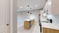 This efficiently designed one-bedroom, one-bathroom unit offers a thoughtful layout across a single floor. The open-concept kitchen serves as the focal point of the main living area, anchored by a substantial center island with a white quartz countertop, an undermount sink, and a built-in dishwasher. Warm light wood cabinetry is paired with white upper cabinets and black hardware, creating a contemporary two-tone aesthetic. Stainless steel appliances, including a refrigerator and range, complete the kitchen. Dual black pendant lights hang above the island, complemented by recessed ceiling lighting throughout. Light-toned laminate flooring runs continuously across the main living area and bedroom, contributing to a clean, cohesive feel. A wall-mounted mini-split unit provides climate control in the main space. The bedroom is a well-proportioned, quiet retreat featuring a large window that frames scenic views of surrounding hills and the neighborhood below, along with a baseboard heater for supplemental warmth. Soft grey walls throughout the unit lend a neutral, modern palette. The bathroom includes a full soaker tub-shower combo with clean white surround. Overall, the unit presents a functional and modern living environment with quality finishes.