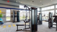 This single-floor fitness center is designed to support a comprehensive workout experience within a spacious, open layout. The main exercise floor is finished with a distinctive patterned carpet featuring bold yellow accent stripes, lending the space an energetic, athletic aesthetic. Walls are decorated with horizontal striping in blue, yellow, and gray tones, reinforcing the facility's dynamic character.

The strength training area is anchored by a series of Matrix cable and selectorized machines, complemented by a full dumbbell rack and a flat utility bench positioned in front of floor-to-ceiling mirrors. A dedicated cardio zone along the perimeter houses a row of treadmills and elliptical trainers, all positioned to face large floor-to-ceiling windows that frame outdoor views of green landscaping and surrounding buildings, filling the space with generous natural light. Multiple ceiling fans and recessed lighting provide consistent airflow and illumination throughout.

A separate, enclosed cycling studio features stationary bikes equipped with individual display screens, set against a deep blue accent wall for an immersive experience. The entry area includes built-in shelving, a small refrigerator, and storage cubbies, along with a glass-enclosed studio space, completing this well-appointed facility.
