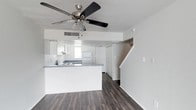 This two-story space offers a clean, functional layout with an open-concept design on the main floor. The living area flows seamlessly into the kitchen, which features a peninsula with a gray countertop — an ideal spot for casual dining or meal prep. White cabinetry lines the kitchen walls, complemented by a classic white refrigerator and a cooktop range. A window above the sink allows natural light to filter into the cooking space, keeping the area bright and airy.

Underfoot, dark hardwood-style flooring extends throughout the main level, providing a rich contrast against the crisp white walls and cabinetry. A ceiling fan with an integrated light fixture and brushed nickel finish anchors the living space overhead, contributing both style and air circulation.

A staircase is visible toward the back of the main floor, leading to the second level. Additional storage is provided by a full-height closet near the base of the stairs. A single light switch panel on the adjacent wall adds to the clean, minimalist aesthetic. The overall ambiance is bright, neutral, and move-in ready.
