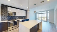 Located at 11560 Old Georgetown Rd in North Bethesda, MD, this single-floor residence offers a thoughtfully designed contemporary living space with a clean, modern aesthetic throughout. The kitchen serves as a standout feature, showcasing a striking dark mosaic tile backsplash that contrasts elegantly with high-gloss white upper cabinetry and deep navy lower cabinets. A large central island with a crisp white quartz countertop provides generous prep and gathering space, illuminated by a pair of industrial-style pendant lights. Stainless steel appliances, including a gas range, built-in microwave, dishwasher, and refrigerator, complete the culinary space. Light hardwood-style flooring flows throughout the main living areas, contributing to a bright and airy atmosphere. The living area benefits from floor-to-ceiling sliding glass doors that open to a balcony, offering elevated outdoor views of the surrounding North Bethesda landscape and flooding the space with natural light. The bedroom features plush carpeting and a large window framing additional outdoor views. The bathroom is finished with large-format gray floor tiles, a soaking tub with an overhead rain showerhead, and a sleek white vanity with a gray countertop, maintaining the residence's cohesive modern character.