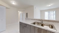 This single-floor space showcases a clean, modern design characterized by bright white walls and large-format light grey porcelain tile flooring that flows throughout the open layout. The kitchen serves as a central feature, equipped with a double stainless steel sink fitted with a sleek chrome faucet. Cabinetry is finished in a contemporary wood-grain grey-brown tone, offering a warm contrast against the light surroundings. The countertops carry a marble-look finish with subtle grey veining, which is echoed in the matching backsplash, creating a cohesive and polished aesthetic. The kitchen peninsula opens directly to the adjacent living area, reinforcing the open-concept design and allowing for easy flow between spaces. Natural light enters through a window visible to the side, brightening the interior. A recessed electrical panel is mounted on the wall nearby, and ceiling-mounted light fixtures provide additional illumination. A hallway visible in the background leads to further rooms, with a white-paneled door offering access to private areas of the unit. Overall, the space presents a fresh, move-in-ready atmosphere with neutral tones and modern finishes throughout.