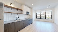 This single-floor unit showcases a thoughtfully designed open-concept layout with a clean, modern aesthetic. Light wood-tone flooring runs continuously throughout the space, complementing the bright white walls and recessed lighting that keep the interior feeling airy and well-lit.

The kitchen is a standout feature, equipped with a full run of dark charcoal lower cabinetry contrasted by crisp white upper cabinets and a gridded white tile backsplash. Open wood shelving with black metal brackets adds a warm, industrial touch, while a matte black gooseneck faucet over a stainless steel sink and a built-in stainless steel microwave complete the functional design. A full-size stainless steel refrigerator anchors one end of the kitchen wall.

The living area is generously proportioned, with large windows framing urban city views and allowing abundant natural light to fill the room. The open floor plan provides flexible arrangement options throughout.

The bathroom features large-format stone-look tile flooring, a soaking tub with shower, matte black fixtures, and a distinctive vertical stack of warm wood-toned niche shelves set into the tiled surround, adding both storage and visual interest to the space.