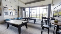 This contemporary amenity space is designed for entertainment and social gatherings, centered around a well-appointed game room that immediately draws the eye. The room features both a pool table and an air hockey table as its focal points, positioned beneath a striking arrangement of modern pendant lights with sleek black-and-gold finishes that add an industrial-chic aesthetic to the space.

Floor-to-ceiling windows spanning an entire wall flood the interior with abundant natural light while offering expansive views of the surrounding outdoor landscape. The open, airy atmosphere created by the high ceilings and generous glazing gives the space a bright and welcoming character.

The flooring consists of a neutral, patterned carpet that ties seamlessly into the room's modern color palette of grays, blacks, and whites, accented by pops of teal from the lounge seating tucked into a casual conversation area along one side. Bar-height seating along the perimeter provides additional spots for spectators or social interaction.

Framed black-and-white artwork adorns the walls, contributing to the sophisticated yet relaxed ambiance. The overall design blends functionality with style, making this an ideal communal gathering space for recreation and leisure.