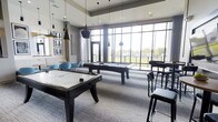 This contemporary amenity space is designed for entertainment and social gatherings, centered around a well-appointed game room that immediately draws the eye. The room features both a pool table and an air hockey table as its focal points, positioned beneath a striking arrangement of modern pendant lights with sleek black-and-gold finishes that add an industrial-chic aesthetic to the space.

Floor-to-ceiling windows spanning an entire wall flood the interior with abundant natural light while offering expansive views of the surrounding outdoor landscape. The open, airy atmosphere created by the high ceilings and generous glazing gives the space a bright and welcoming character.

The flooring consists of a neutral, patterned carpet that ties seamlessly into the room's modern color palette of grays, blacks, and whites, accented by pops of teal from the lounge seating tucked into a casual conversation area along one side. Bar-height seating along the perimeter provides additional spots for spectators or social interaction.

Framed black-and-white artwork adorns the walls, contributing to the sophisticated yet relaxed ambiance. The overall design blends functionality with style, making this an ideal communal gathering space for recreation and leisure.