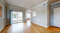 This thoughtfully designed multi-level unit is accessed via a carpeted staircase fitted with a warm wood handrail and flanked by neutral-toned textured walls, with a window at the top lending natural light to the entry. The main living area is bright and open, featuring gleaming hardwood floors, crown molding, and a sliding glass door that opens to a private balcony with views of the surrounding neighborhood. A built-in shelving nook and a pass-through counter add functional architectural character to the living and dining spaces. The kitchen is well-equipped, showcasing rich dark wood cabinetry, black appliances including a refrigerator, range, and over-the-range microwave, a double sink with ample counter space, and the same hardwood flooring that flows throughout the main level. The bedrooms are generously sized, finished with plush carpeting, ceiling fans, and large windows that invite natural light. One bedroom includes access to a walk-in closet. The bathroom is well-appointed with a wide double-sink vanity topped with a light stone surface, dark wood cabinetry, a tiled shower-tub combo, and hardwood flooring. An in-unit laundry area is also visible, adding everyday convenience to this comfortable and functional home.