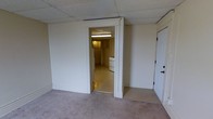 This single-floor space presents a compact and utilitarian layout well-suited for practical use. The main room features wall-to-wall carpeting and smooth white walls, creating a neutral and clean backdrop. A drop tile ceiling runs throughout, giving the space a consistent, low-profile overhead finish. Fluorescent lighting provides steady illumination across the interior. An interior doorway connects the main room to a separate kitchen area, which is finished with hard flooring and includes a white gas range, offering a dedicated space for cooking and meal preparation. Cabinetry lines the kitchen walls, providing functional storage. A solid white entry door with hardware is positioned in the main room, serving as the primary point of access. The overall character of the space is straightforward and move-in ready, with a no-frills design that prioritizes function. The consistent white wall finish throughout unifies the rooms, while the separation between living and kitchen areas adds a degree of defined organization to the floor plan.
