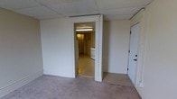 This single-floor space presents a compact and utilitarian layout well-suited for practical use. The main room features wall-to-wall carpeting and smooth white walls, creating a neutral and clean backdrop. A drop tile ceiling runs throughout, giving the space a consistent, low-profile overhead finish. Fluorescent lighting provides steady illumination across the interior. An interior doorway connects the main room to a separate kitchen area, which is finished with hard flooring and includes a white gas range, offering a dedicated space for cooking and meal preparation. Cabinetry lines the kitchen walls, providing functional storage. A solid white entry door with hardware is positioned in the main room, serving as the primary point of access. The overall character of the space is straightforward and move-in ready, with a no-frills design that prioritizes function. The consistent white wall finish throughout unifies the rooms, while the separation between living and kitchen areas adds a degree of defined organization to the floor plan.