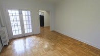 This single-floor residence offers a clean, functional layout with a welcoming open feel. The main living area is finished with classic parquet hardwood flooring in a warm, light wood tone, adding visual texture and character to the space. Crisp white walls and white baseboards throughout create a bright, neutral backdrop that complements a variety of interior styles. A French door with multi-pane glass panels allows natural light to filter into the living area, enhancing the sense of openness. The living area flows seamlessly into the kitchen through an open doorway, allowing for easy movement between spaces. The kitchen is visible from the main room and features a black refrigerator, with cabinetry providing practical storage. The layout is straightforward and well-proportioned, making efficient use of the available space. The overall ambiance is simple and understated, offering a solid foundation for personalization. The consistent flooring and neutral color palette unify the space, giving it a cohesive and tidy appearance throughout.