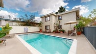 This inviting outdoor space centers around a generously sized rectangular swimming pool with clear turquoise water, accessible via stainless steel handrails and surrounded by a wide concrete pool deck. The deck is thoughtfully furnished for both relaxation and outdoor dining, featuring multiple reclining sun loungers positioned along the poolside for sunbathing, as well as a shaded dining area equipped with a table, chairs, and a large patio umbrella. The surrounding building is constructed in a classic stucco style with a neutral beige exterior, dark brown-trimmed windows, and flat rooflines accented in places by traditional clay tile detailing. The complex wraps around the pool area, creating an enclosed, courtyard-like setting that enhances privacy and a sense of community. Abundant tropical and subtropical plantings — including banana trees, flowering shrubs, and large potted plants — add vibrant greenery and color throughout the space. A perimeter fence with iron railings separates portions of the pool area from adjacent building entryways. The open-air setting benefits from natural daylight, with mature trees visible beyond the roofline, contributing to a relaxed, resort-like atmosphere well-suited for outdoor recreation and leisure.