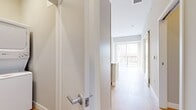 This single-floor, one-bedroom, two-bathroom unit offers a thoughtfully designed layout that maximizes its compact footprint. The open-concept kitchen is a standout feature, showcasing sage-toned shaker-style cabinetry with matte black hardware, quartz countertops with subtle veining, a white subway tile backsplash, and a full suite of stainless steel appliances including a dishwasher, range, microwave, and refrigerator. A peninsula provides additional counter space and separates the kitchen from the living area. Pendant lighting adds a modern touch overhead. Light-toned hardwood flooring runs throughout the main living areas, contributing to a bright, cohesive aesthetic. A dedicated hallway leads to the bedroom, which features plush carpeting, a large window, and a sliding glass door opening to a private balcony with open outdoor views. Two bathrooms include white vanities with matte black fixtures and a tub-shower combination. A convenient in-unit stacked washer and dryer is tucked into a dedicated closet. Neutral warm-gray walls throughout give the space a clean, contemporary feel.