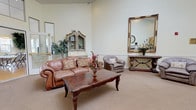 This single-floor community space is designed to provide comfort and functionality across a variety of thoughtfully arranged areas. The welcoming lounge areas feature warm, neutral tones, plush seating including leather sofas and upholstered armchairs, ornate mirrors with decorative console tables, and carpeted flooring that adds to the inviting ambiance. Crown molding and recessed lighting enhance the refined character of these gathering spaces.

The fully equipped kitchen is outfitted with ample oak cabinetry, a full suite of appliances including a range, refrigerator, and microwave, and a generous island with bar-height seating — ideal for group meals or social gatherings. The adjacent dining areas feature light hardwood flooring and multiple windows dressed with sheer curtains and cheerful yellow accent valances, allowing abundant natural light to fill each room.

The spacious activity room boasts elevated ceilings, wide windows, and an open hardwood floor plan well-suited for events, meetings, or group activities. Flexible folding tables and chairs allow the space to be configured as needed. Wide, arched hallways connect the various wings of the facility, maintaining an open and navigable flow throughout. The overall atmosphere is warm, welcoming, and purposefully designed for community use.