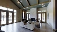 This single-story space makes an immediate impression with its dramatic vaulted ceiling, where dark-painted exposed beams contrast elegantly against the coffered panels above. The ceiling height creates an expansive, airy atmosphere throughout the main living area. A floor-to-ceiling fireplace serves as the room's focal centerpiece, clad in a bold chevron-patterned tile in neutral tones that draws the eye upward and anchors the seating arrangement below. The living area is furnished with a mix of cream upholstered sofas and striped accent chairs, arranged to encourage conversation while maintaining an open, uncluttered feel. Wide-plank hardwood-style flooring in a cool gray tone runs throughout, complementing the warm, earthy wall colors. Natural light floods the space through multiple sets of glass French doors and large windows fitted with layered curtains in deep brown and gold tones. These doors provide direct access to outdoor areas on multiple sides, enhancing the sense of connection to the exterior. Recessed lighting supplements the natural light, ensuring the space remains well-lit throughout the day. The overall ambiance is refined yet welcoming, blending contemporary design elements with transitional furnishings.