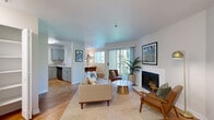 This thoughtfully designed 2-bedroom, 1-bathroom residence offers a cohesive layout across a single floor, with rooms that flow naturally from one to the next. The open-concept living area is anchored by a white tile-surround wood-burning fireplace and complemented by warm hardwood flooring, a neutral color palette, and a sliding glass door that fills the space with natural light and views of surrounding greenery. The adjacent dining area features a bright window overlooking lush trees, creating an inviting atmosphere for everyday meals. The kitchen is efficiently laid out with gray shaker cabinetry, light quartz countertops, a stainless steel sink, and includes a dishwasher and electric range — providing practical functionality in a clean, modern aesthetic. Both bedrooms are carpeted and tranquil, each offering closet space and natural light; the primary bedroom is comfortably sized for a queen bed and furnishings. A hallway connects the bedrooms to the shared full bathroom, which includes a tub-shower combo and hardwood-style flooring. An entrance area and hallway complete the layout, providing organized transitions between spaces. Overall, the residence presents a bright, mid-century influenced aesthetic with functional room distribution and a warm, livable character throughout.