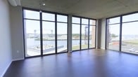 This single-floor space offers an impressive open-plan layout defined by architectural clarity and industrial refinement. The most striking feature is the expansive wall of floor-to-ceiling windows framed in dark metal, which floods the interior with natural light and frames unobstructed panoramic views of a nearby waterway and an iconic lift bridge beyond. The exposed concrete ceiling adds a raw, industrial character that contrasts with the smooth, polished concrete flooring underfoot, creating a cohesive modern aesthetic throughout. A structural concrete column punctuates the window line, complementing the building's honest use of materials. An adjacent brick wall element introduces warmth and texture to the otherwise cool palette. The open, column-free layout provides maximum flexibility for a variety of uses. Outside, a terrace with seating is visible just below, further emphasizing the connection to the waterfront setting. The overall ambiance is one of openness and urban sophistication, combining generous natural light, industrial materials, and compelling views into a highly functional and visually dynamic space.