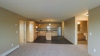 This single-floor space is designed with an open-concept layout that seamlessly connects the kitchen and living areas, creating a cohesive and functional environment. The kitchen is a standout feature, showcasing light wood cabinetry that extends to the upper walls, offering generous storage throughout. A central island provides additional workspace and serves as a natural focal point, complemented by a stylish pendant light hanging overhead. Stainless steel appliances, including a refrigerator and range, add a modern touch to the warm, neutral cabinetry tones. Dark flooring in the kitchen zone creates a clear visual distinction from the adjacent living area, which is finished with wall-to-wall carpet for a comfortable, inviting feel. The neutral color palette of warm beige and taupe tones carries throughout the space, lending a calm and cohesive ambiance. The open sightlines between the kitchen and living area make the space feel expansive and bright. Additional doorways visible from the main living zone suggest further rooms or utility spaces beyond, enhancing the overall functionality of this well-organized, single-level layout.