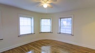 This single-floor space showcases a distinctive curved room that immediately draws the eye with its graceful, rounded walls — a classic architectural detail that adds character and charm. Three generously sized double-hung windows fitted with horizontal blinds line the curved wall, flooding the room with an abundance of natural light and offering views of the surrounding neighborhood. The windows are framed with clean white molding that complements the bright, neutral wall tones throughout.

Underfoot, warm hardwood flooring runs the length of the room, its polished surface reflecting the incoming light and contributing to the airy, open feel of the space. White baseboard trim adds a crisp, finished look along the base of the curved walls. Overhead, a white ceiling fan with a built-in light fixture provides both ambient lighting and practical ventilation, enhancing everyday comfort.

The overall ambiance is clean, bright, and welcoming, with a timeless quality rooted in its classic architectural details. The curved room serves as a standout feature, offering a flexible and inviting area suitable for a variety of uses. The combination of natural light, hardwood floors, and thoughtful design details makes this a well-appointed and functional single-floor space.