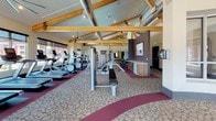 This fitness center offers a spacious and well-appointed workout environment defined by its striking architectural character. The vaulted ceiling features prominently exposed wooden beams alongside visible industrial ductwork, creating a dynamic aesthetic that balances rustic warmth with modern functionality. Ceiling fans are positioned throughout to maintain comfortable airflow during workouts.

The facility is generously illuminated by natural light streaming through large, floor-to-ceiling windows along multiple walls, providing open views of the surrounding community. This abundance of glazing gives the space an airy, energizing atmosphere well-suited for exercise.

The main workout floor is finished with patterned carpet accented by bold burgundy borders, lending the space a structured yet inviting feel. The cardio zone features multiple treadmills and elliptical machines aligned along the windowed walls, while the strength training area houses a full cable machine with bench, a selection of Precor selectorized machines, and a dumbbell rack. Large mirrors further enhance the sense of depth throughout.

Near the entrance, a water fountain station is set against a textured mosaic accent wall, accompanied by a glass-enclosed vestibule. The overall layout is open and well-organized, offering users a comprehensive and comfortable training environment.