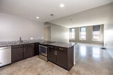 This single-floor unit is defined by a clean, contemporary design with a neutral color palette and an open layout that allows the kitchen and living areas to flow seamlessly together. The kitchen is well-appointed with dark espresso cabinetry, granite countertops featuring subtle speckled patterning, and a full suite of stainless steel appliances including a range, refrigerator, microwave, and dishwasher. A peninsula with an extended granite surface serves as a functional divider between the kitchen and the expansive living area, which benefits from natural light filtering through multiple windows. Recessed lighting throughout provides consistent illumination. Speckled terrazzo-style flooring runs continuously across the main living areas, hallways, and bedrooms, contributing to the cohesive aesthetic. The hallway connects several private rooms, each accessed through dark-paneled interior doors that add a modern contrast to the bright white walls. A walk-in closet is outfitted with adjustable wire shelving systems, maximizing storage capacity. The bathroom features a granite-topped vanity with an undermount sink and a generously sized walk-in tiled shower with built-in shelving and a bench seat.