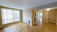 This single-floor space offers a clean, functional layout highlighted by warm-toned hardwood flooring that runs throughout. The main living area is bright and open, benefiting from a large window dressed with vertical blinds that filters generous natural light into the room. A classic white radiator sits beneath the window, providing an efficient heating solution. The neutral, beige-toned walls create a warm and inviting ambiance that complements the natural wood flooring.

A built-in storage unit with mirrored sliding doors adds a practical organizational feature while enhancing the sense of space. A hallway connects the living area to additional rooms, one of which is visible in the background and similarly benefits from natural light through its own window. The flow between spaces is open and unobstructed, lending the overall layout a spacious feel.

The space is well-maintained and move-in ready, with clean white trim and baseboards providing a crisp contrast to the warmer wall tones. The combination of hardwood floors, ample natural light, and a straightforward layout makes this a versatile and welcoming space suitable for a variety of uses.