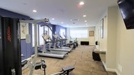 This fitness center offers a thoughtfully designed workout space spread across a single floor. The room features a clean, modern aesthetic with a combination of bold blue and neutral beige walls that create an energizing yet comfortable atmosphere. Recessed lighting throughout the ceiling ensures the space is well-lit, while multiple windows with white shutters allow natural light to supplement the bright interior.

The cardio area is prominently equipped with a row of treadmills and an elliptical trainer, all featuring integrated display consoles for tracking workout metrics. Strength training is supported by a multi-function cable machine situated at the front of the room, along with a weight bench for targeted exercises. The flooring consists of durable, patterned carpet tiles that provide comfort and stability underfoot during workouts.

Along the perimeter, a water fountain and a small counter area offer convenient hydration and storage solutions. A large wall mirror enhances the sense of space and allows users to monitor their form during exercise. Framed artwork adds a finishing touch to the walls, contributing to a polished and welcoming environment. The overall layout is open and functional, providing ample room to move freely between equipment while maintaining an organized and tidy appearance.