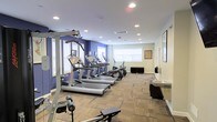 This fitness center offers a thoughtfully designed workout space spread across a single floor. The room features a clean, modern aesthetic with a combination of bold blue and neutral beige walls that create an energizing yet comfortable atmosphere. Recessed lighting throughout the ceiling ensures the space is well-lit, while multiple windows with white shutters allow natural light to supplement the bright interior.

The cardio area is prominently equipped with a row of treadmills and an elliptical trainer, all featuring integrated display consoles for tracking workout metrics. Strength training is supported by a multi-function cable machine situated at the front of the room, along with a weight bench for targeted exercises. The flooring consists of durable, patterned carpet tiles that provide comfort and stability underfoot during workouts.

Along the perimeter, a water fountain and a small counter area offer convenient hydration and storage solutions. A large wall mirror enhances the sense of space and allows users to monitor their form during exercise. Framed artwork adds a finishing touch to the walls, contributing to a polished and welcoming environment. The overall layout is open and functional, providing ample room to move freely between equipment while maintaining an organized and tidy appearance.