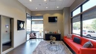 This single-floor space presents a well-appointed lounge or reception area designed with a contemporary aesthetic. The room is anchored by a bold orange sofa adorned with accent pillows, complemented by a sleek brown armchair, creating a welcoming and visually dynamic seating arrangement. A plush, light-toned shag area rug grounds the space, while a minimalist glass and gold coffee table adds an elegant focal point.

The walls are finished in a warm neutral tone, providing a clean backdrop for the wall-mounted flat-screen television positioned above a dark, low-profile credenza. The credenza serves as both a functional and decorative element, displaying carefully curated accessories including colorful vases and sculptural accents.

Natural light flows generously into the space through large floor-to-ceiling windows and a glass-paneled entrance, offering views of the surrounding exterior. Recessed lighting in the drop ceiling supplements the natural light, ensuring the interior remains bright and inviting. Dark-toned flooring throughout adds a grounding contrast to the lighter furnishings and walls, contributing to the space's modern, polished ambiance.