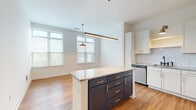 Located at 96 E Catawba St in Belmont, NC, this thoughtfully designed one-bedroom, one-bathroom unit offers a efficient and stylish layout. The open-concept kitchen and living area is a standout feature, anchored by a two-toned center island with dark lower cabinetry, gold hardware, and a granite countertop. White shaker-style upper cabinets, a classic subway tile backsplash, and stainless steel appliances — including a French door refrigerator, electric range, and built-in microwave — complete the kitchen. Pendant and linear lighting fixtures illuminate the space, while multiple windows allow generous natural light to fill the open living area. Warm hardwood-style flooring runs throughout the entire unit, adding continuity and warmth. A clean, well-lit hallway connects the main living area to the private quarters, and features a smart home panel for added convenience. The bedroom is a comfortable size with clean white walls and ample room for furnishings. Adjacent to the bedroom is a generously sized walk-in closet outfitted with built-in shelving and double hanging rods. The bathroom is elegantly finished with a granite-topped vanity, black fixture accents, gold cabinet hardware, and a full tub-shower combination surrounded by white tile.