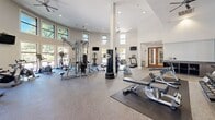 Located at 431 El Camino Real in Santa Clara, CA, this well-appointed fitness center offers a generously sized, single-floor exercise space designed to accommodate a wide range of workout needs. The facility is characterized by soaring ceilings that create an open, airy atmosphere, enhanced by rows of large, elevated windows that flood the interior with natural light and frame views of the surrounding palm-lined landscape. Ceiling fans are strategically positioned throughout to ensure comfortable air circulation during workouts.

The exercise room is organized into distinct functional zones. A dedicated cardio area features multiple treadmills, elliptical machines, stationary and spin bikes, and a stair climber, all arranged to maximize space and usability. A robust strength training section includes a multi-station cable machine, free weights, dumbbells, medicine balls, and adjustable benches. A large floor-to-ceiling mirror along one accent wall visually expands the space and supports form monitoring.

The facility is accessed through warm wood-framed French doors, lending an inviting architectural detail to the entrance. Durable, textured flooring covers the exercise area, providing both safety and comfort. Wall-mounted flat-screen televisions are positioned throughout the space for entertainment during workouts. The overall design balances functionality with a clean, modern aesthetic.
