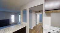This single-floor residence offers a well-designed open layout that connects the main living areas with ease and functionality. The kitchen is a standout feature, showcasing sleek granite countertops with a distinctive veined pattern, dark cabinetry, and a built-in sink — all complemented by a textured ceiling that adds subtle character throughout the space. Structural columns define the transition between rooms while maintaining an open, airy feel.

The flooring shifts thoughtfully between warm hardwood-style planks in the kitchen and hallway areas and plush carpet in the living room, providing both comfort and visual variety. Natural light filters into the living area through a glass door or window, brightening the neutral, soft gray and white color palette that is carried consistently throughout the space.

A central hallway connects the main living areas to the additional rooms, with multiple doors providing access to what appear to be bedrooms, offering privacy while maintaining a cohesive flow. HVAC vents are integrated into the walls and ceilings, ensuring climate comfort throughout. The clean lines, neutral tones, and practical finishes make this a versatile and move-in-ready space well-suited for a variety of lifestyles.