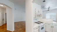 This single-floor residence presents a clean, cohesive design anchored by warm hardwood-style flooring that runs throughout the entire space, complementing the crisp white walls and trim. The layout flows naturally from room to room, with arched architectural details adding a refined touch to the transitions between spaces.

The kitchen is well-appointed and functional, featuring white cabinetry with brushed nickel hardware, white quartz-style countertops, a gas range with overhead microwave, and a dishwasher. An arched passageway leads from the kitchen into the adjoining living areas, enhancing the open feel of the floor plan.

The living area offers generous proportions with bright, neutral walls and ample natural light filtering through window blinds. The spacious bedroom includes a walk-in closet, providing practical storage, and is served by two bathrooms. The primary bathroom is outfitted with a single-sink vanity featuring white cabinetry, a large mirror with Hollywood-style lighting, and a full soaking tub with white tile surround. A second bathroom mirrors this clean aesthetic with a tub-shower combination and matching white finishes. The hallway connecting the rooms is wide and airy, reinforcing the open and uncluttered character of the home throughout.