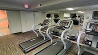 This fitness center is a dedicated, single-floor exercise space designed to support an effective cardio workout routine. The room is outfitted with multiple Star Trac treadmills, each equipped with individual touchscreen monitors, allowing users to track metrics or enjoy entertainment during their workout. The mirrored walls that line the space serve a dual purpose: they visually expand the room and allow users to monitor their form while exercising. The patterned carpet flooring adds a layer of texture and comfort underfoot, complementing the functional aesthetic of the space. Overhead lighting provides consistent, even illumination throughout the room, ensuring a well-lit environment at any time of day. A clearly marked emergency exit door enhances safety, reflecting a thoughtful approach to the layout. The overall ambiance is clean, utilitarian, and purpose-driven, offering everything needed for a focused cardiovascular training session. This fitness space is well-suited for individuals seeking a convenient and properly maintained workout facility.