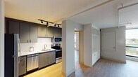 This single-floor unit showcases a contemporary design with clean lines, neutral tones, and light hardwood flooring that flows throughout most of the space. The kitchen is well-appointed with dark grey cabinetry, a full suite of stainless steel appliances including a refrigerator, electric range, microwave, and dishwasher, complemented by a white countertop and a stainless steel sink. Track lighting overhead provides focused illumination, adding a modern industrial touch to the cooking area. A structural dividing wall separates the kitchen from the adjacent living area, which features built-in white cabinetry offering additional storage and display space.

The bedroom is bright and airy, benefiting from a large floor-to-ceiling window that frames outdoor city views and allows abundant natural light. A sliding closet door reveals an organized interior with shelving and a built-in dresser.

The bathroom is finished with a grey vanity topped with a white surface, an illuminated round mirror, and a full tub-shower combination with white tile surround. A dedicated laundry area houses a stacked washer and dryer unit, providing convenient in-unit functionality. The overall ambiance is clean, functional, and modern.