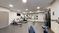 This fitness room offers a well-rounded exercise environment designed for both cardio and strength training. The space is finished with a durable speckled rubber floor, smooth white walls, and a clean white ceiling fitted with recessed pot lighting that ensures the room is consistently and evenly illuminated throughout.

Cardio equipment is prominently featured and includes a treadmill, elliptical trainer, rowing machine, and stationary bikes from Matrix, providing a wide variety of aerobic workout options. For strength training, the room is equipped with a multi-station cable machine, a dumbbell rack with a full set of free weights ranging across multiple sizes, and an adjustable workout bench. A wall-mounted flat-screen television is positioned for viewing during exercise sessions.

A large mirror spans a significant portion of one wall, visually expanding the room and allowing users to monitor their form during workouts. A split-system air conditioning unit is mounted on the wall, ensuring comfortable climate control year-round. Natural light filters in through a window offering a view of surrounding greenery, adding a refreshing element to the workout environment. Access is provided through a solid double door as well as a secondary door leading to an adjacent corridor. The overall ambiance is modern, clean, and functional.