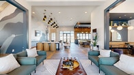 Located at 110 1 Mile Parkway in Nashville, TN, this single-floor community clubhouse delivers a sophisticated, open-plan environment thoughtfully organized into multiple distinct yet seamlessly connected zones. The main lounge area commands attention with soaring ceilings accented by a bold teal-painted tray ceiling detail and a striking gold chandelier, complemented by teal upholstered sofas, a tufted leather ottoman, and a large-scale abstract mural that anchors the space with artistic flair. The flooring throughout features an eye-catching pattern of large-format gray tiles interspersed with warm wood inlays, adding visual depth and guiding the flow between areas. A dedicated dining and bar zone showcases warm wood-paneled walls, a dark hexagonal tile floor, and a bold yellow island beneath a row of gold diamond-shaped pendant lights, all framed by expansive windows that invite abundant natural light and exterior views. A separate intimate lounge nook features a deep teal accent ceiling, arched backlit mirrors, built-in banquette seating, and a wet bar with marble countertops — ideal for private gatherings. Curated gallery walls, designer pendant clusters, and carefully selected furnishings throughout reflect a cohesive, eclectic aesthetic that balances modern elegance with approachable comfort.