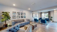 Located at 7200 N Frontage Rd in Orlando, FL, this thoughtfully designed single-floor amenity space offers a sophisticated and welcoming environment for residents and guests alike. The lounge is organized into distinct yet flowing zones, creating a versatile layout suited for both relaxed socializing and casual gatherings.

The main seating area showcases tufted gray sofas and armchairs arranged around glass-topped coffee tables, complemented by a curated gallery of framed antique-style mirrors that add depth and elegance to the light gray walls. Recessed lighting and large-format tile flooring contribute to the polished, modern aesthetic throughout.

A secondary seating nook features vibrant teal velvet armchairs centered around a hexagonal marble coffee table, with framed line-art prints depicting wine motifs adorning the walls — adding a playful, upscale character to the space. Patterned area rugs and patterned drapery panels frame floor-to-ceiling windows that flood the room with natural light and offer open views of the exterior grounds.

The kitchenette area is appointed with sleek gray cabinetry, matte black hardware, white countertops, and glass-front upper cabinets, providing a refined and functional space for entertaining. High-top bistro tables with tufted leather seating near the large windows create an additional casual dining area. Glass partition walls with black frames elegantly define zones while maintaining an open, airy feel throughout.