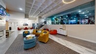 Located at 1000 Elm St in Manchester, NH, this generously sized amenity floor offers a diverse collection of thoughtfully designed spaces catering to fitness, leisure, and community. The welcoming lobby lounge serves as the central hub, featuring polished marble-style tile flooring, a coffered ceiling, and an eclectic mix of colorful seating — including a curved mustard velvet sofa, teal accent chairs, and bean bag chairs — alongside a striking digital display wall and a convenience retail area. A glass-enclosed library nook provides a quiet retreat lined with white bookshelves and cozy armchairs. The expansive fitness center is well-appointed with a full suite of strength and cardio equipment, including cable machines, power racks, barbells, and treadmills, all set on durable rubber flooring with glass partitions and accent teal columns. A dedicated corridor connects to a glass-walled screening room outfitted with stadium-style seating and ambient blue LED lighting. The game room features hardwood-style flooring, a pool table, ping-pong table, arcade games, and bold teal accent walls. A large event and dining space accommodates group gatherings with long communal tables, stylish blue and gold seating, and elegant chandelier lighting throughout.
