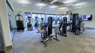 This fitness center offers a fully outfitted workout environment designed to support a wide range of exercise routines. The space is anchored by an extensive collection of strength training machines, including cable machines, chest press stations, and multi-use resistance equipment, all arranged to allow comfortable movement between stations. Along the perimeter, a row of cardio machines — including ellipticals, stair climbers, and treadmills — lines the wall of large windows, allowing natural light to flood the room and providing views of the surrounding outdoor greenery. The generous ceiling height, fitted with rows of linear fluorescent lighting, contributes to a bright and energizing atmosphere throughout the day. Neutral-toned carpet tiles with a subtle striped pattern cover the floor, adding both comfort underfoot and a clean, modern aesthetic. The open layout ensures ample space between equipment, promoting an uncluttered and accessible environment for users of all fitness levels. Overall, the fitness center is a thoughtfully designed, functional space that balances practicality with a welcoming ambiance suited for daily training.
