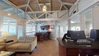 Located in Hoover, Alabama, this single-story community clubhouse at 3627 Cedarbrook Drive is defined by its striking architectural character and welcoming open layout. The centerpiece of the space is a soaring vaulted ceiling with exposed white-painted truss work and warm wood planking, lending the interior a grand yet inviting atmosphere. Large clerestory windows wrap the upper walls, flooding the main lounge area with natural light and offering views of the surrounding outdoor areas, including what appears to be a screened porch and fitness space beyond.

The main lounge features comfortable tufted neutral sofas, a glass-top coffee table, and a fireplace set into a dark wood cabinet along a slate-blue accent wall, creating a cozy gathering area. A wall-mounted flat-screen television adds to the functionality of this common space. Adjacent to the lounge is a reception and business area with a dark granite-topped counter and computer workstations, ideal for resident services.

The kitchenette is well-appointed with rich dark wood cabinetry, granite countertops, a stainless steel microwave, and a double-basin sink — providing practical amenities for events and daily use. Throughout, large-format tile flooring ties the spaces together cohesively, while track lighting and ceiling fans ensure comfort and visibility across the entire facility.