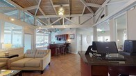 Located in Hoover, Alabama, this single-story community clubhouse at 3627 Cedarbrook Drive is defined by its striking architectural character and welcoming open layout. The centerpiece of the space is a soaring vaulted ceiling with exposed white-painted truss work and warm wood planking, lending the interior a grand yet inviting atmosphere. Large clerestory windows wrap the upper walls, flooding the main lounge area with natural light and offering views of the surrounding outdoor areas, including what appears to be a screened porch and fitness space beyond.

The main lounge features comfortable tufted neutral sofas, a glass-top coffee table, and a fireplace set into a dark wood cabinet along a slate-blue accent wall, creating a cozy gathering area. A wall-mounted flat-screen television adds to the functionality of this common space. Adjacent to the lounge is a reception and business area with a dark granite-topped counter and computer workstations, ideal for resident services.

The kitchenette is well-appointed with rich dark wood cabinetry, granite countertops, a stainless steel microwave, and a double-basin sink — providing practical amenities for events and daily use. Throughout, large-format tile flooring ties the spaces together cohesively, while track lighting and ceiling fans ensure comfort and visibility across the entire facility.