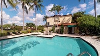 This Mediterranean-inspired residential community presents a striking outdoor environment defined by its warm stucco exteriors, terracotta tile roofing, and richly landscaped surroundings. The centerpiece of the property is a generously sized swimming pool with crystal-clear turquoise water, set against a backdrop of soaring palm trees that frame the sky and lend the space a distinctly tropical character. The pool deck is finished with elegant stone pavers and features a seating area with outdoor furniture, ideal for relaxation and socializing. A pergola-style structure anchors one corner of the outdoor space, adding architectural interest and shade. Adjacent to the pool, a cabana or recreational structure with a green awning and French doors provides additional amenity space. The building itself rises to two stories, with balconies and large windows that look out over the lush grounds. A decorative wrought-iron gate with ornamental tile accents marks the entry to the pool area, offering both security and visual charm. Mature tropical trees and carefully tended plantings with potted greenery line the perimeter, creating a private, resort-like atmosphere. The overall ambiance is one of relaxed, sun-drenched outdoor living rooted in classic Mediterranean design.